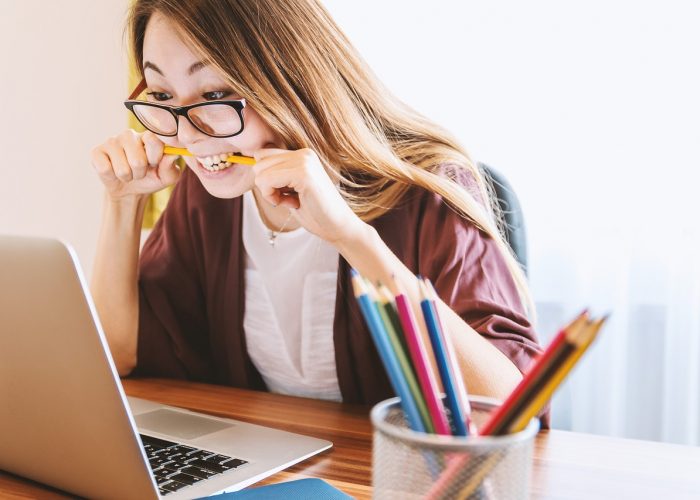 Flicka med långt hår och glasögon som sitter framför en bärbar dator och har en penna i munnen.