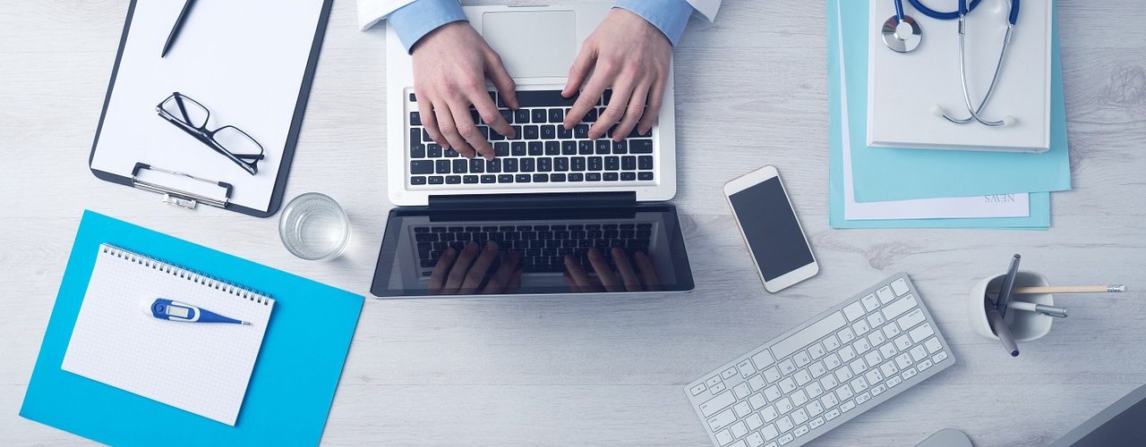 Läkare som sitter vid sin dator. Stetoskop och andra verktyg ligger runt honom.